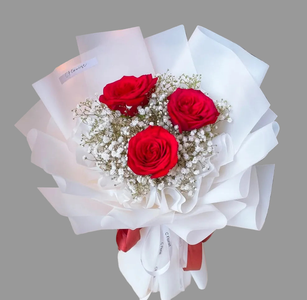 A bouquet of red roses with baby's breath and white floral paper, tied with a ribbon handle and adorned with a bow.