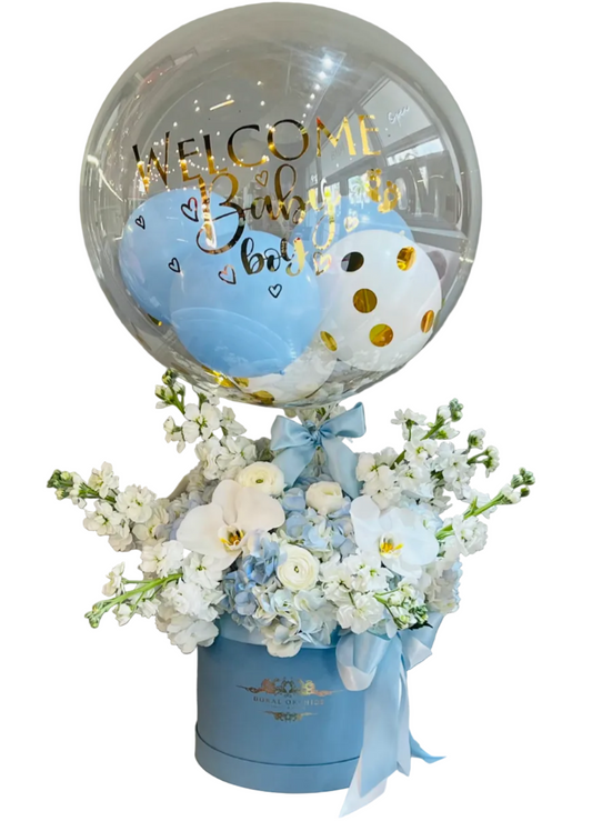 Decorative arrangement with a transparent balloon featuring 'Welcome Baby Boy' text, surrounded by flowers in a blue box.